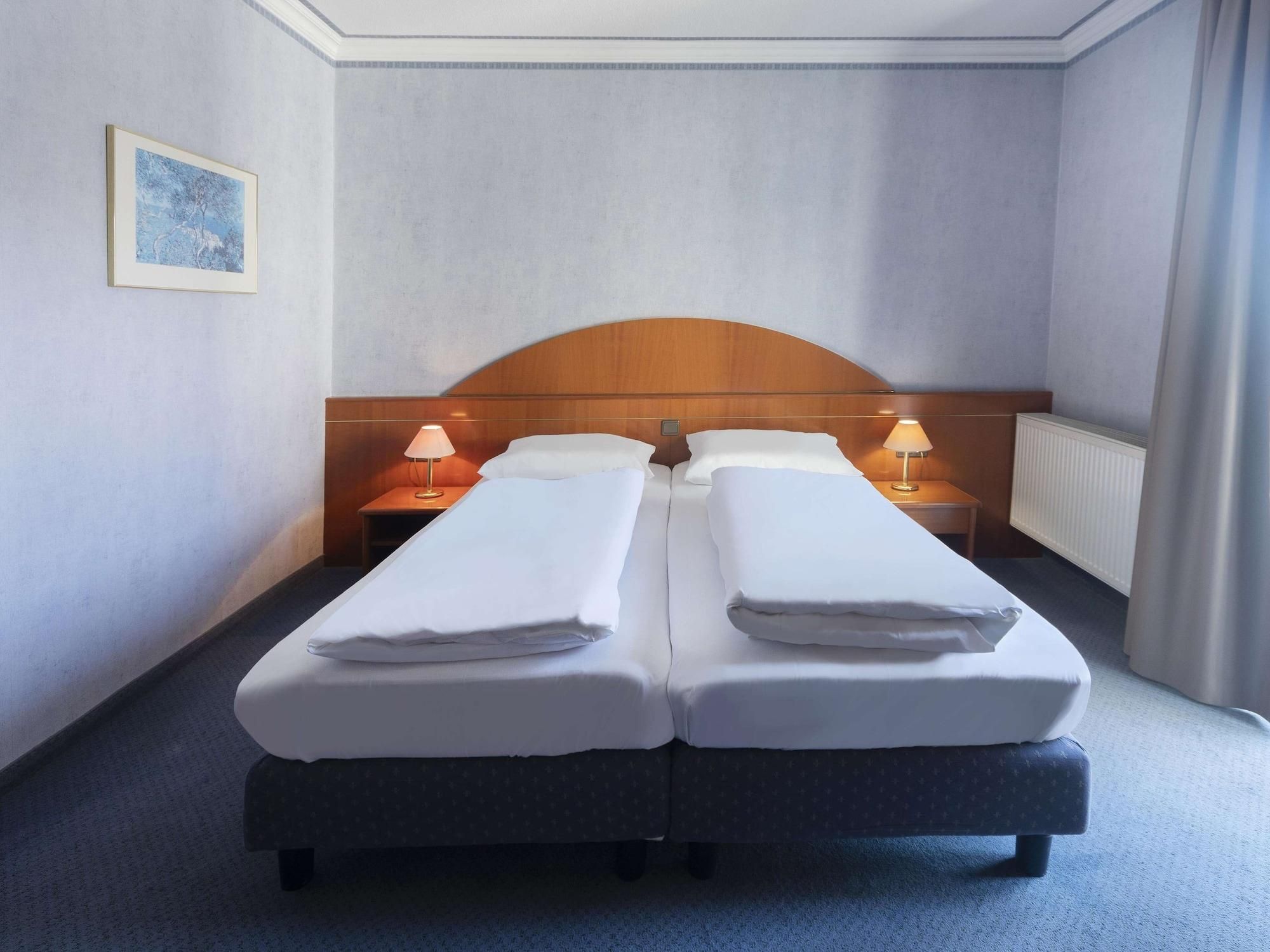 Hotellihuone, jossa on kaksi erillistä sänkyä, puinen pääty, yöpöytävalaisimet, patteri ja seinällä oleva kehystetty kuva.
