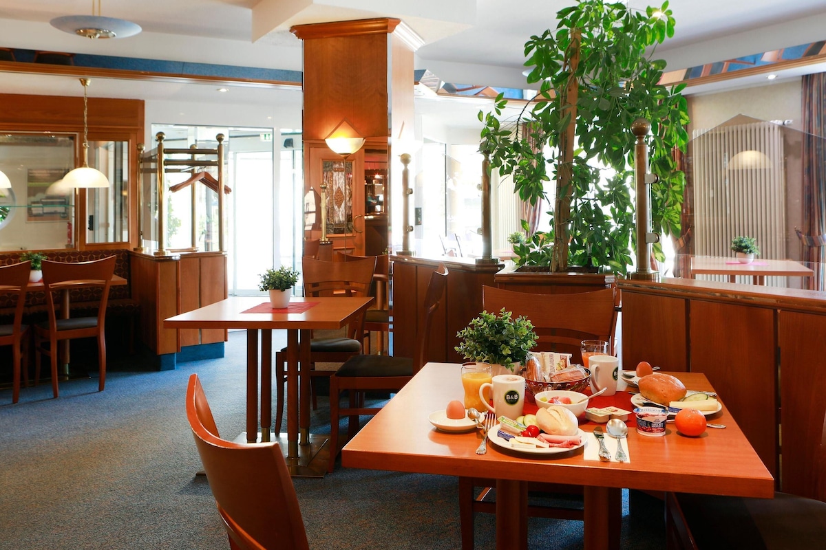 A dining area with wooden tables and chairs, a large potted plant, and a view of the street outside.