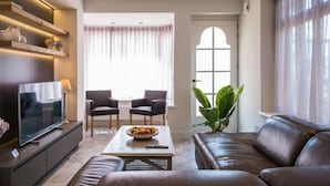 Un salon moderne doté d’un téléviseur à écran plat, de deux fauteuils, d’une table basse avec des fruits, d’un canapé et d’une grande fenêtre avec des rideaux.
