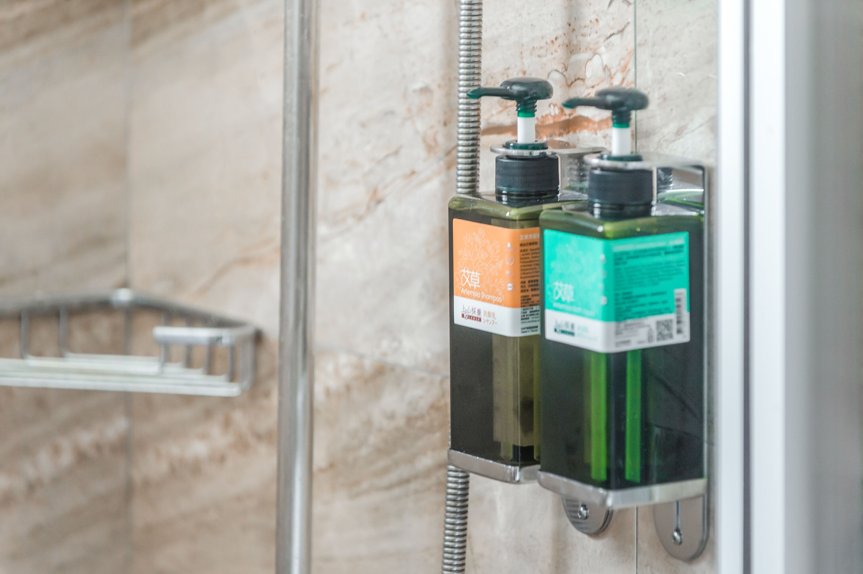 A bathroom with a marble wall and a glass shower enclosure. There are two bottles of shampoo and conditioner on a shelf.