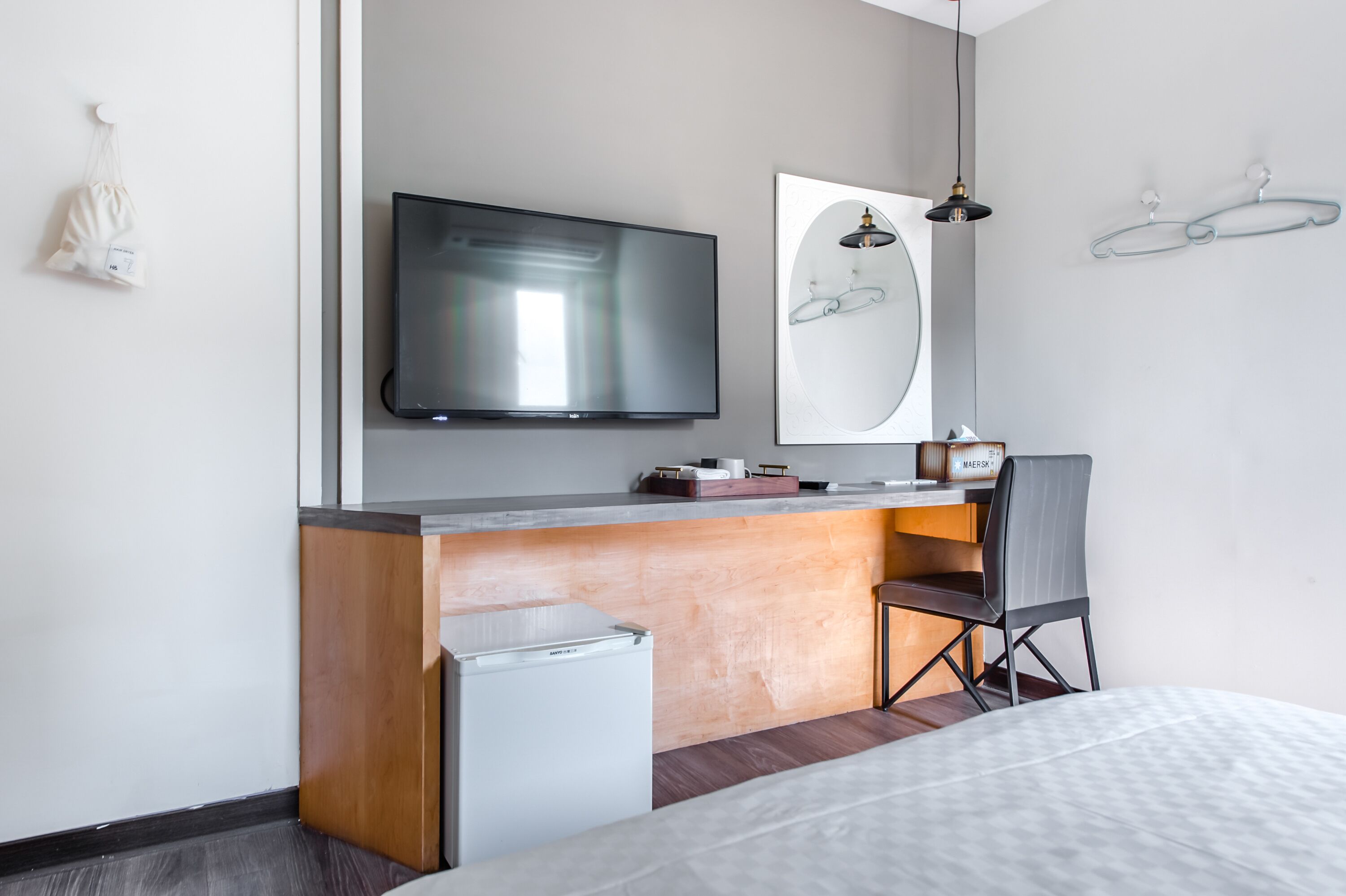 Une chambre d’hôtel moderne dotée d’un téléviseur à écran plat fixé au mur, d’un bureau en bois avec une chaise, d’un petit réfrigérateur et d’un lit recouvert d’une couverture blanche à texture.