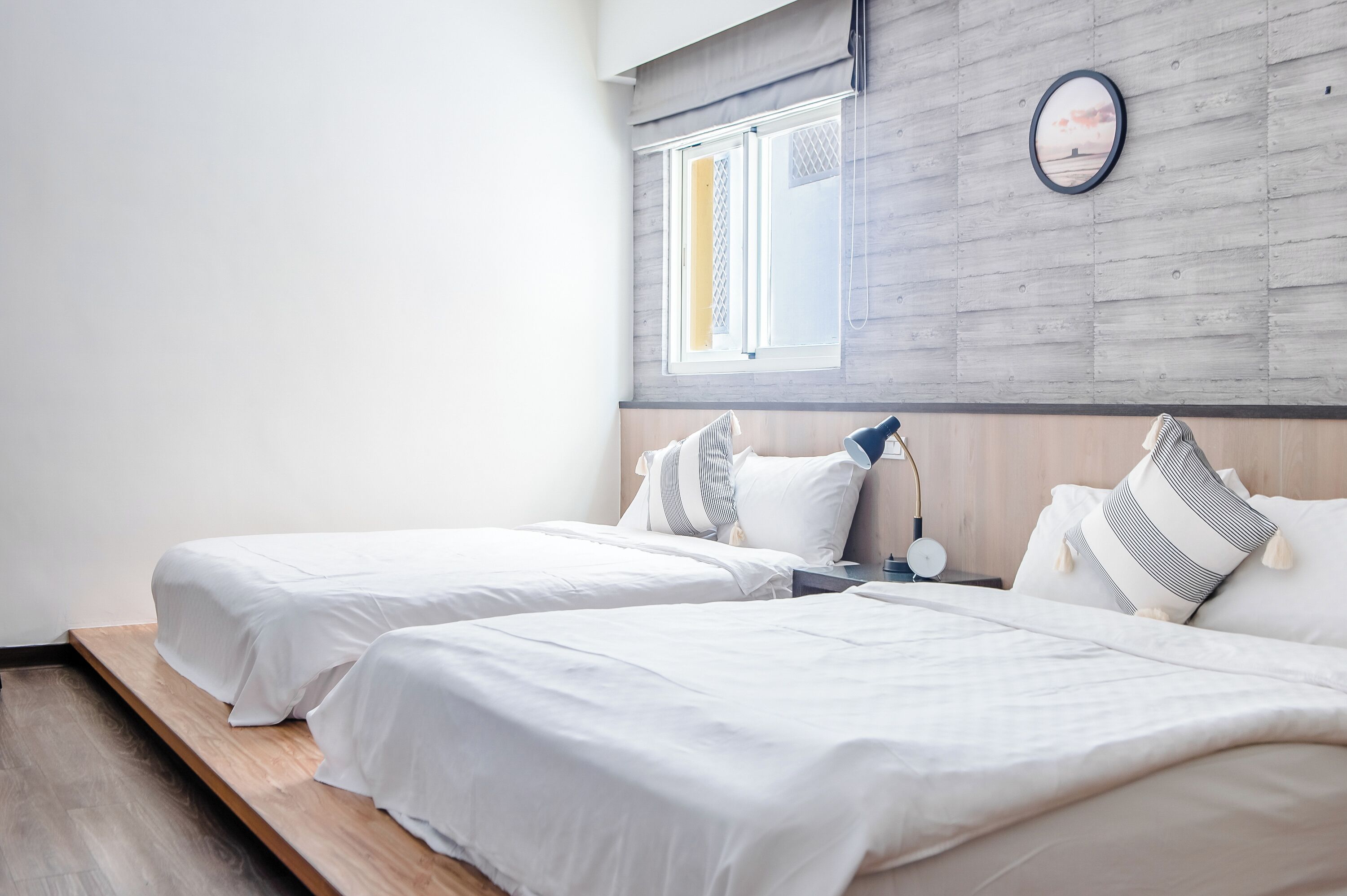 A hotel room with two beds, a wooden floor, a window with blinds, and a clock on the wall.