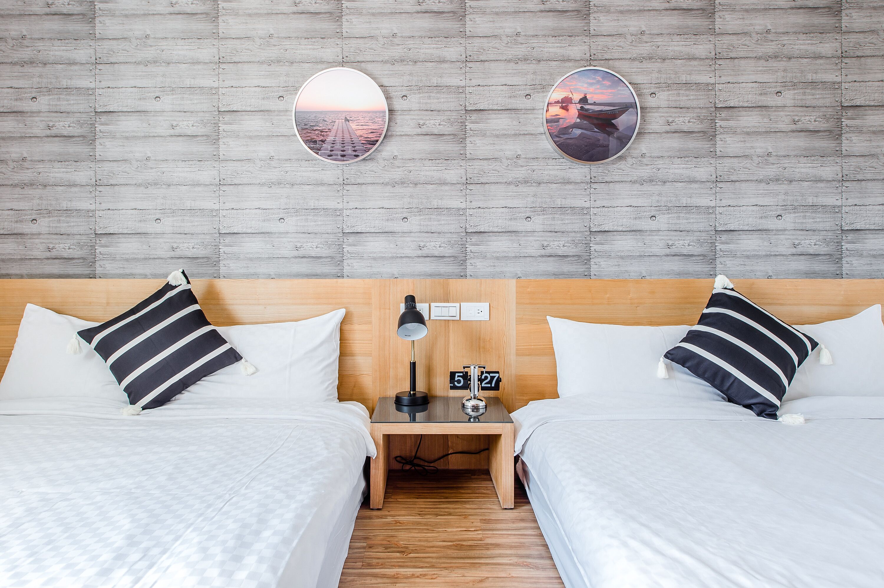 Habitación de hotel con dos camas, cabecera de madera, mesita de noche con un reloj y dos cuadros colgados en la pared.