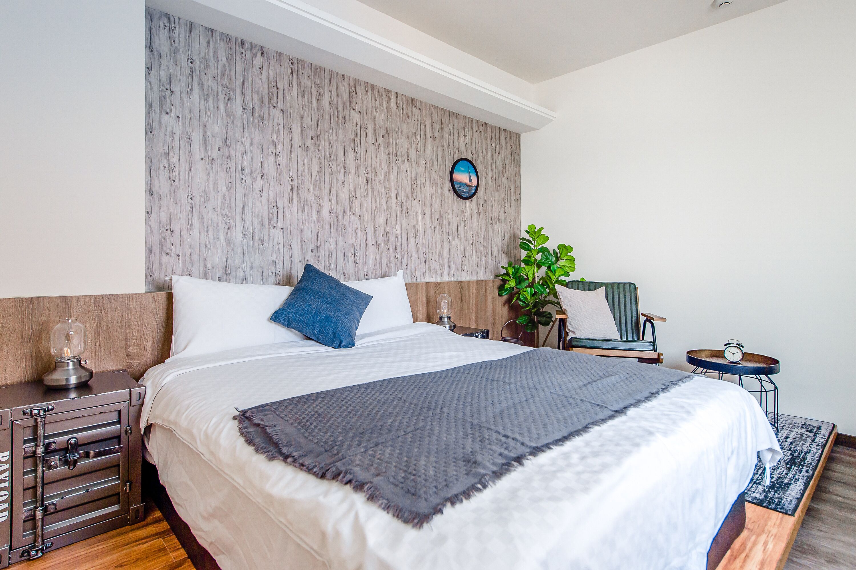 A neatly made bed with a white and gray blanket, a blue pillow, and a wooden headboard. There is a small table with a clock and a plant beside the bed.