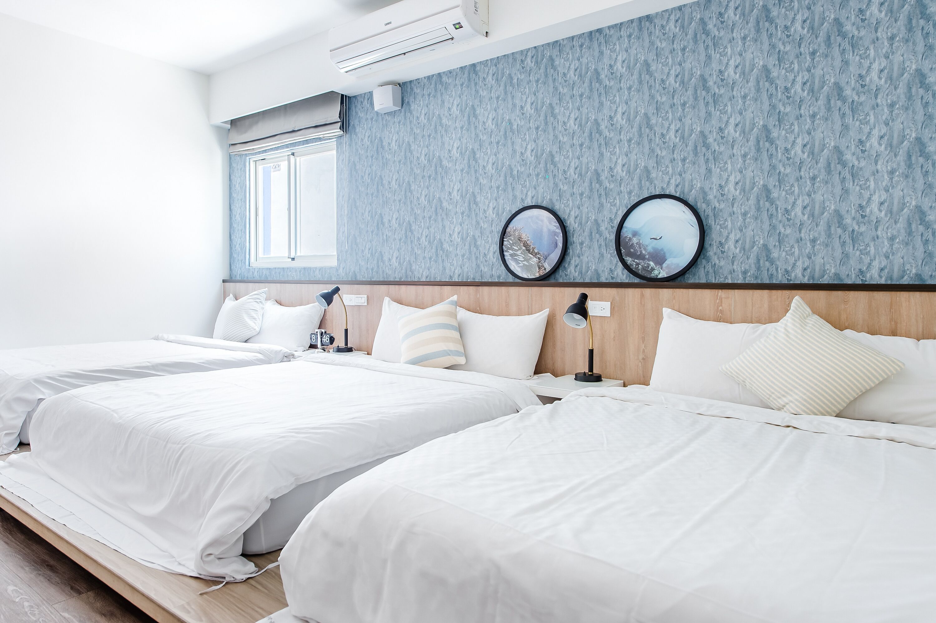 Habitación de hotel con dos camas, cabecero de madera, ventana con persianas y dos cuadros colgados en la pared.