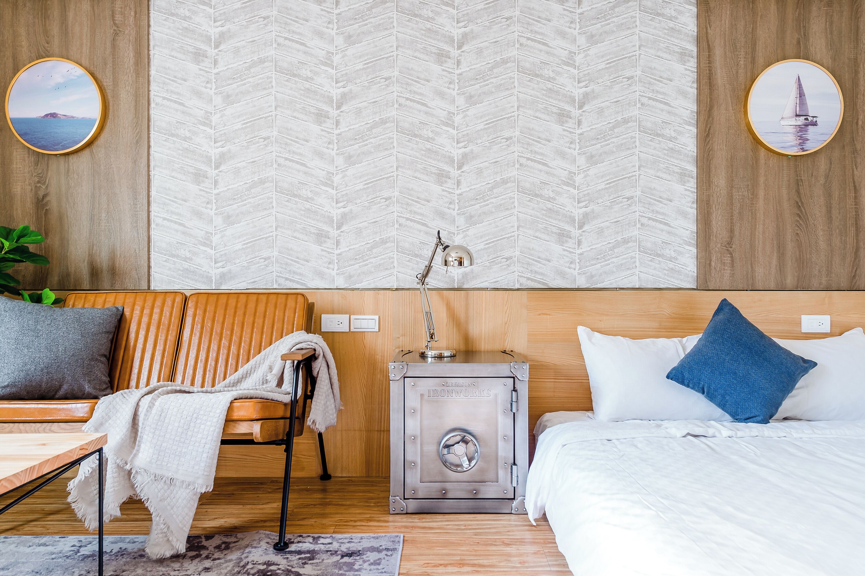A modern hotel room with a herringbone-patterned wallpaper, a wooden headboard, a bed with white bedding, a small bedside table, a lamp, and a safe.
