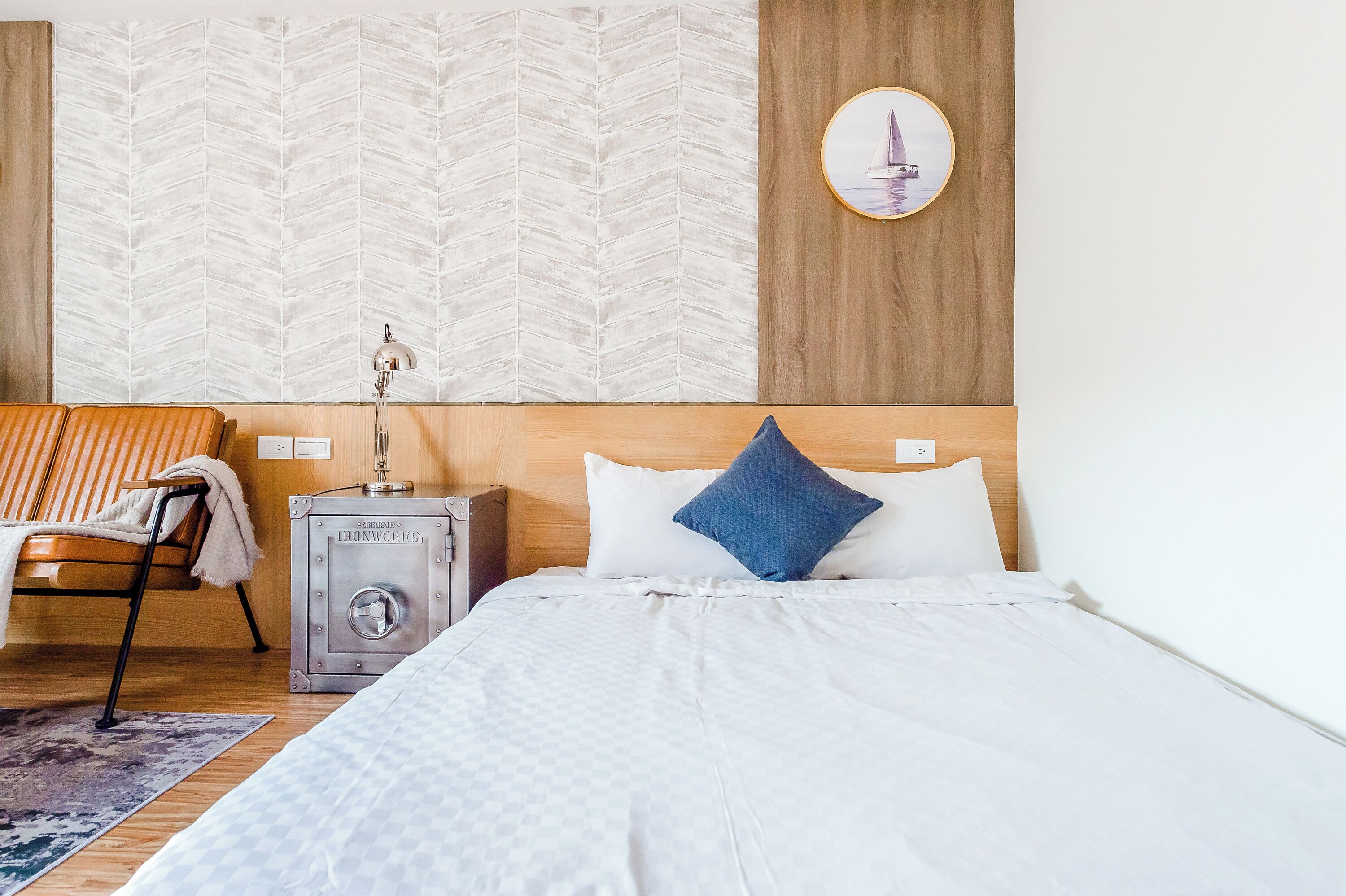 A hotel room with a bed, a nightstand, a lamp, a chair, and a wall with a leaf-patterned wallpaper.