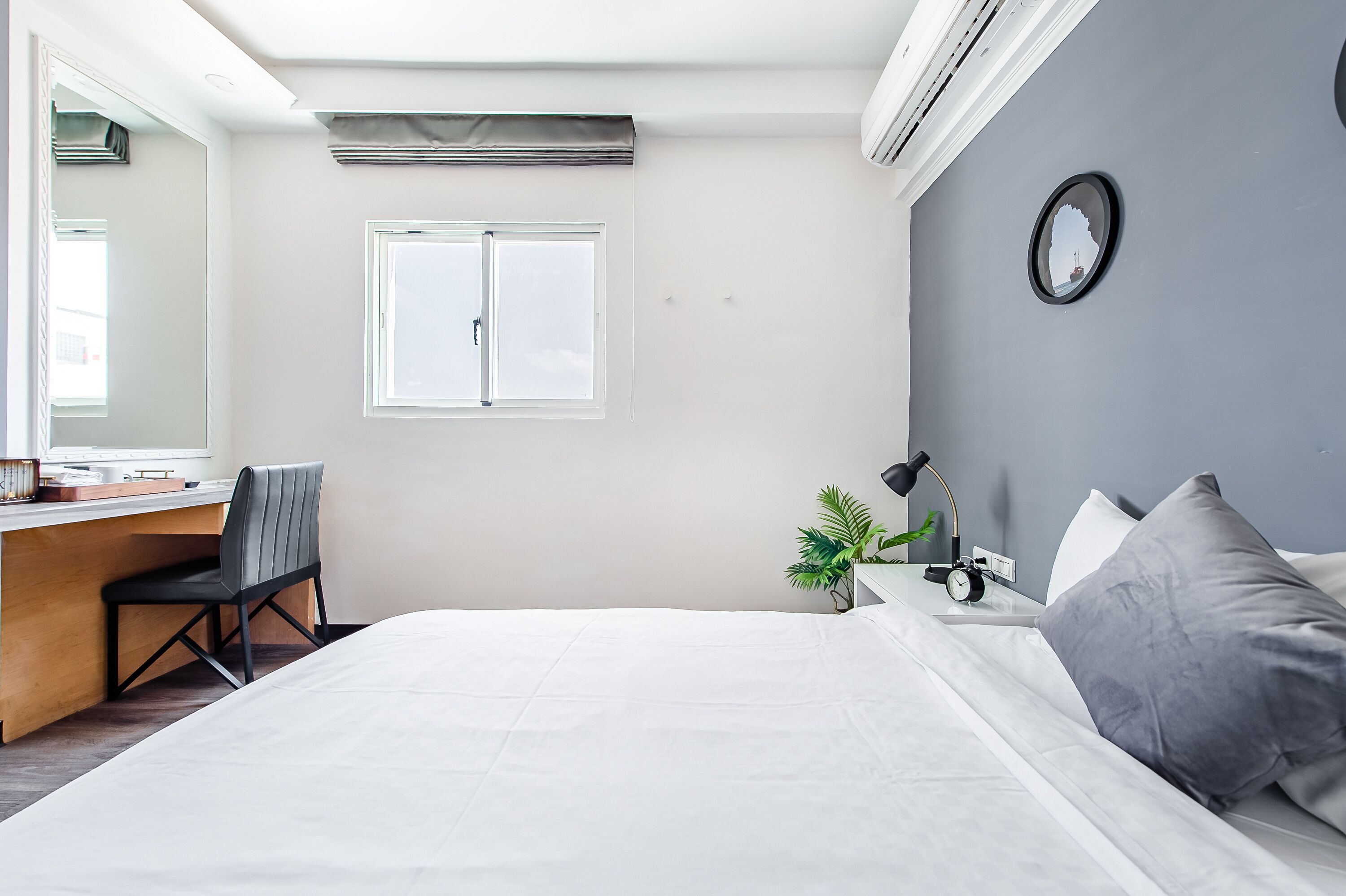 Un dormitorio con cama, escritorio, silla, planta pequeña, reloj y ventana.