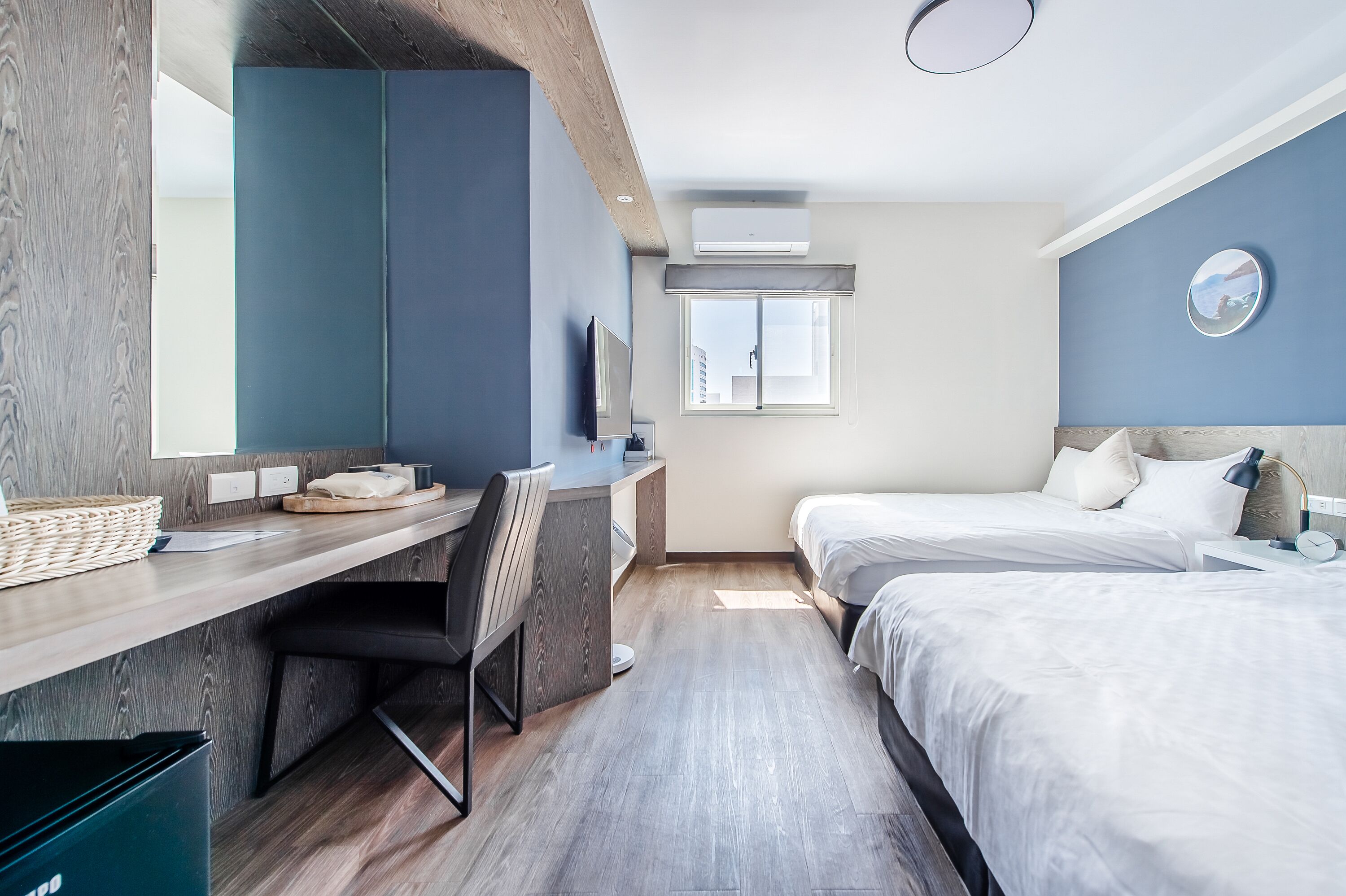 A modern hotel room with a wooden desk, a bed, and a large window.