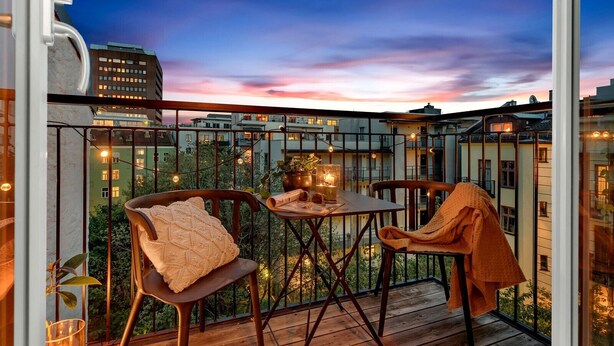 Un balcon avec une table et des chaises, des guirlandes lumineuses et une vue sur les bâtiments au crépuscule.