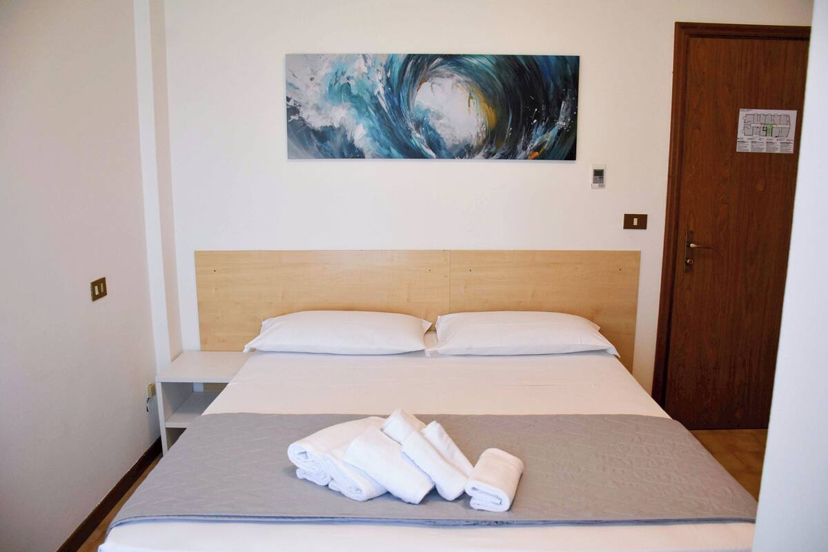 A hotel room with a bed, a painting on the wall, and a door with a sign.