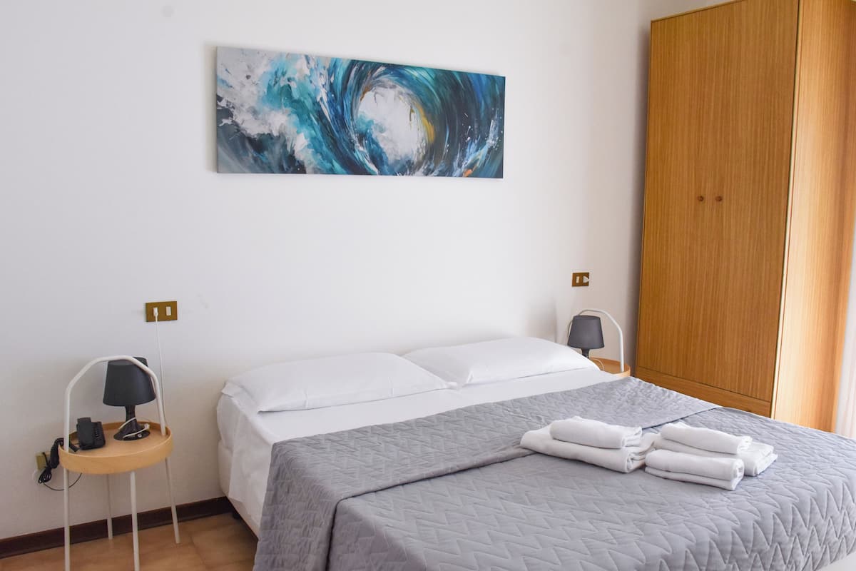 A hotel room with a bed, a desk with a lamp, a wooden wardrobe, and a painting on the wall.