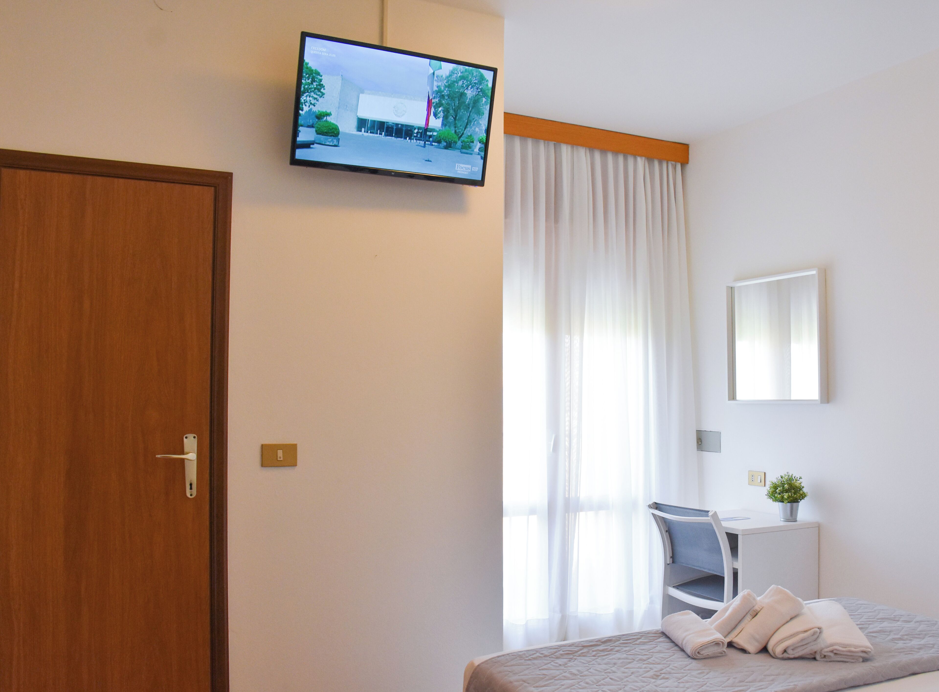 A hotel room with a wooden door, a bed with a quilted cover, a desk with a chair, a television mounted on the wall, and a window with white curtains.