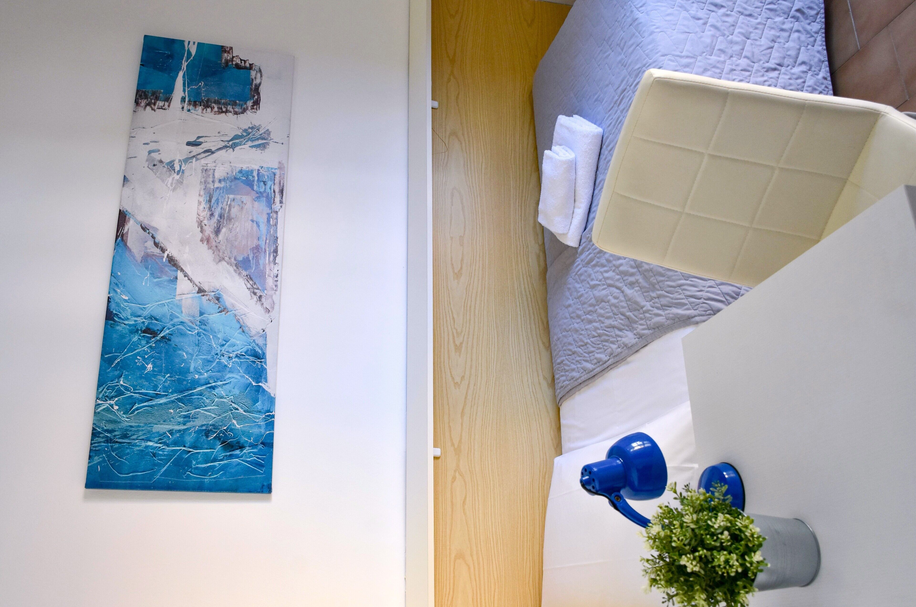 Ein Zimmer mit einem Bett, einem an der Wand hängenden Kunstwerk, einem Nachttisch mit einer blauen Lampe und einer kleinen Pflanze.