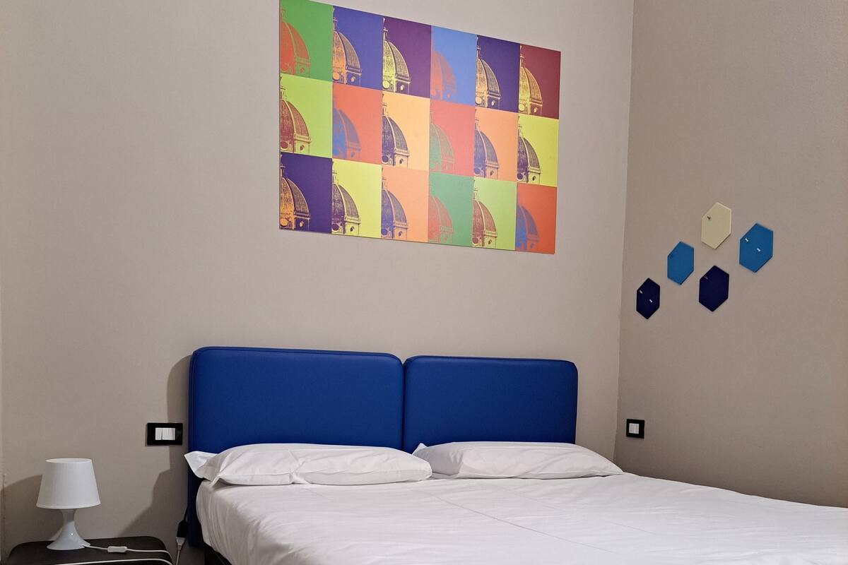 A bedroom with a bed, a nightstand, a lamp, a colorful wall art, and hexagonal wall decorations.