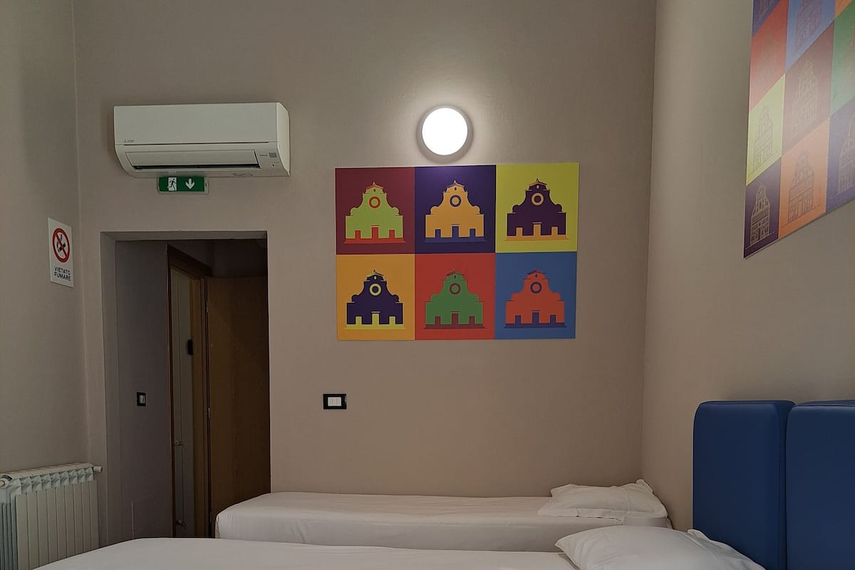 A hotel room with a large bed, a wall-mounted air conditioner, a door, and a colorful wall decoration.