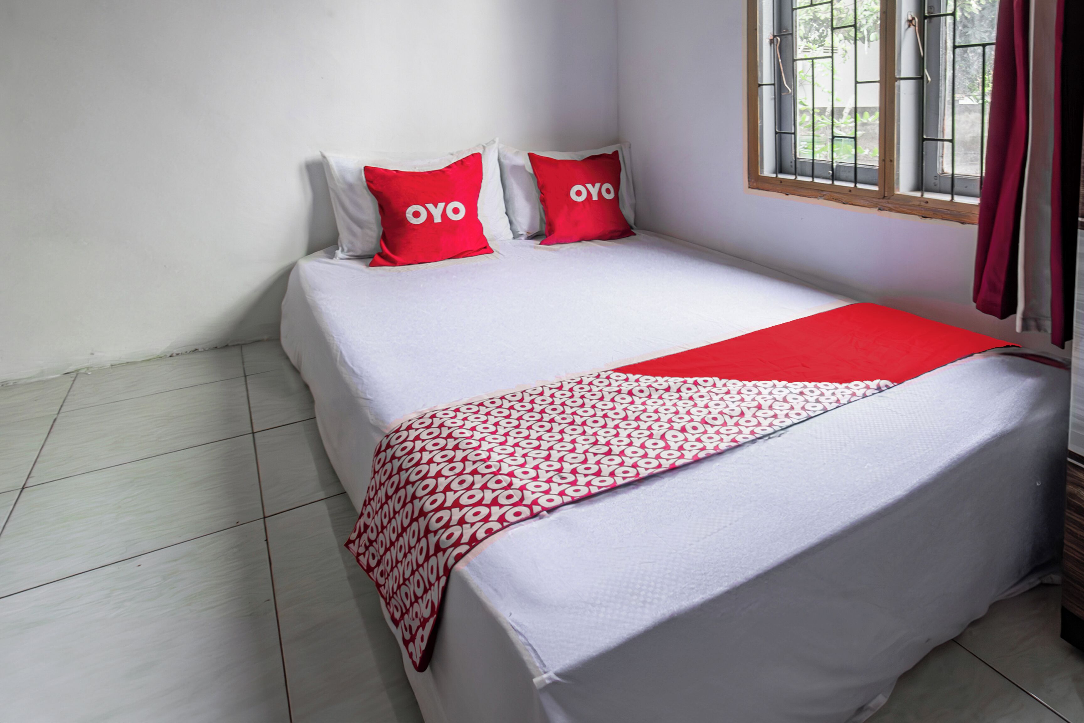 Une chambre avec un lit simple, recouverte d’une courtepointe blanche, agrémentée de deux oreillers rouges portant le logo « OYO », d’une couverture rouge à motifs blancs et d’une fenêtre avec un grillage métallique.