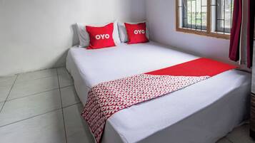 Une chambre avec un lit simple, recouverte d’une courtepointe blanche, agrémentée de deux oreillers rouges portant le logo « OYO », d’une couverture rouge à motifs blancs et d’une fenêtre avec un grillage métallique.
