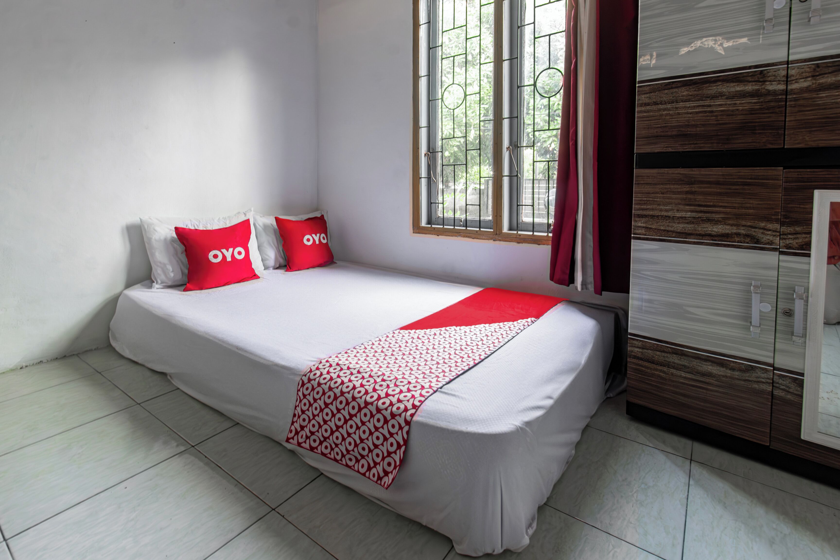 A single bed with a white mattress and red accents, a window with metal bars, and a wooden wardrobe.