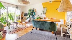 A modern living room with a wooden dining table, a green sofa, and a yellow accent wall. - Large Two-bedroom Flat to Rent in Paris,100m From Beautiful Buttes Chaumont Parc (Paris)