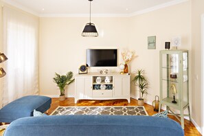 A modern living room with a flat-screen TV, a white entertainment unit, two blue sofas, a patterned rug, and a green cabinet with glass doors.