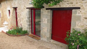 A stone building with a red door and windows, a small garden with flowers, and a white chair outside. - Gîte Écoconçu Charmant Proche zoo de la Flèche : Idéal Famille (CLERMONT CREANS)