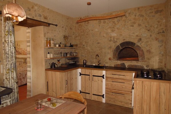 A rustic kitchen with wooden cabinets, a stone wall, a built-in oven, and a dining area with a table and chairs.