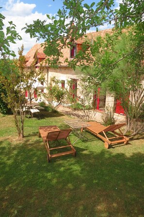 A garden with wooden lounge chairs and a table, surrounded by greenery and a building with red accents.