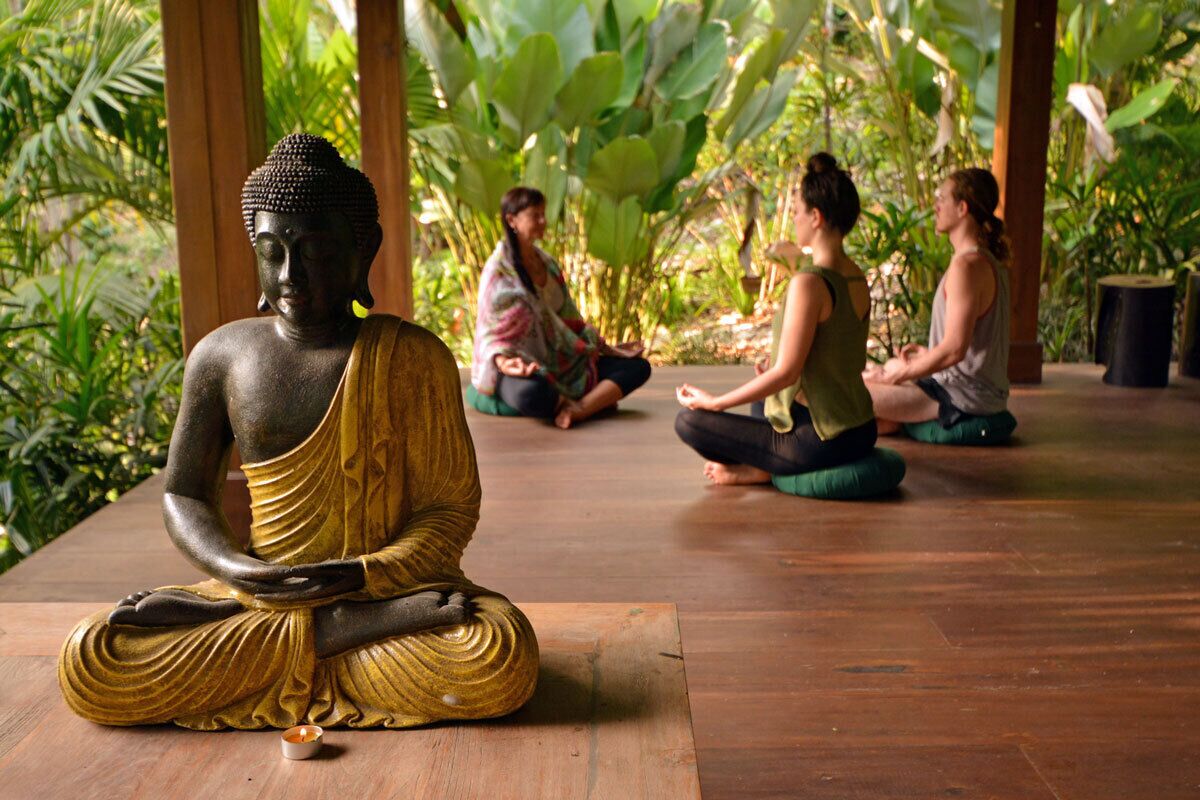 A Buddha statue in a meditative pose with three people sitting in a circle in the background.
