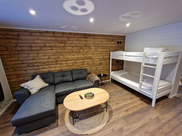 Una habitación con pared de madera, un sofá gris, una cama litera blanca y una mesa de centro redonda con un control remoto y una planta pequeña.
