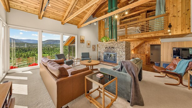 A spacious living room with a high ceiling, a stone fireplace, and large windows offering a view of the outdoors. - Cliff Cabin | Hot Tub | Spectacular Views (Fairmont Hot Springs)