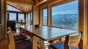 A wooden interior with a large table and chairs, large windows offering a mountain view. - Cliff Cabin | Hot Tub | Spectacular Views (Fairmont Hot Springs)