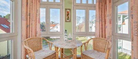 A room with two wicker chairs around a marble table, large windows, and a view of the outside.