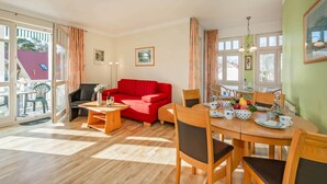 A spacious living area with a red sofa, wooden dining table, and chairs, a small side table with a vase and wine glasses, and a balcony with a view of a red-roofed building. - Cozy vacation apartment by the sea with balcony, WiFi & parking space (Koserow)