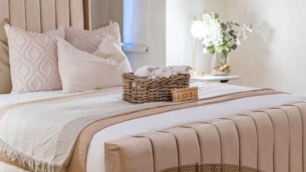 A neatly made bed with a woven basket containing folded towels, a bedside table with a lamp, and a vase with flowers.