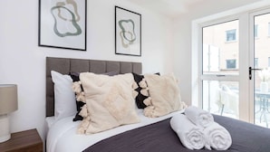 A neatly made bed with a grey headboard, two framed abstract artworks, and a pair of rolled towels on the bed.