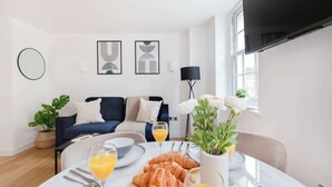 A modern living room with a dining table set for breakfast, featuring croissants and orange juice. There is a sofa with cushions, a potted plant, and artwork on the wall.
