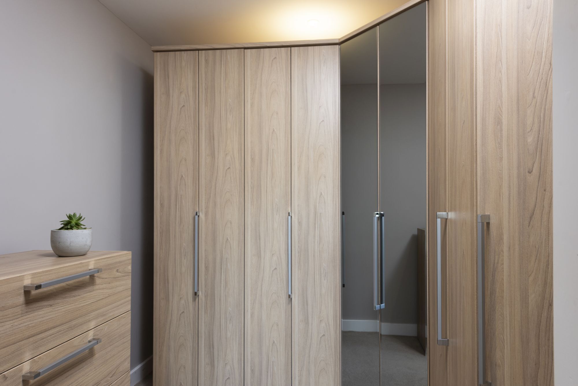 Ein Zimmer mit einem Holzschrank, einer kleinen Pflanze auf einem Schminktisch und einem Spiegel an der Wand.