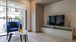 A modern living room with a large window, a blue armchair, a small table, and a TV on a white cabinet. - w* | Homey 3BR with Balcony in Premium Miraflores (Miraflores)