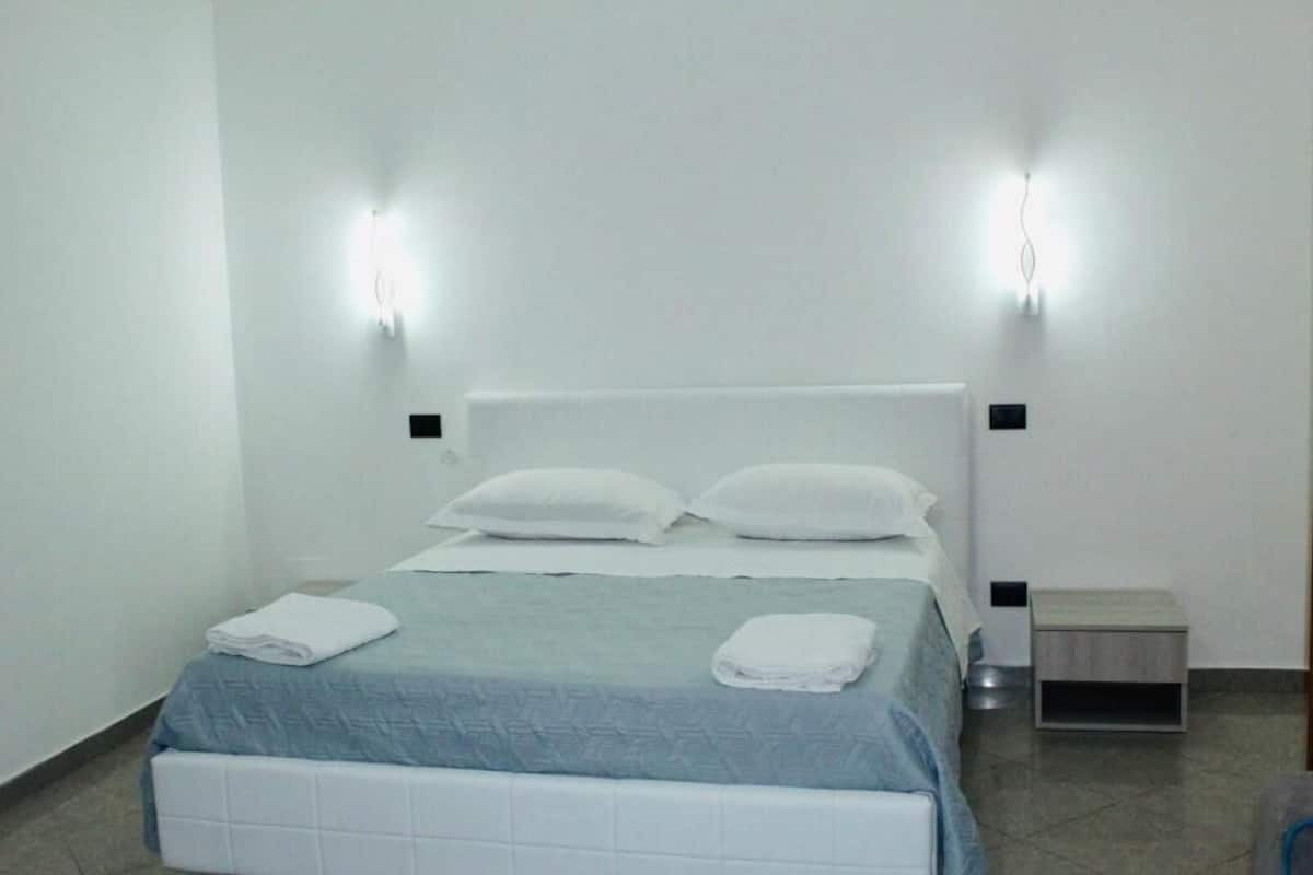 A clean, modern hotel room with a bed, two bedside tables, and wall-mounted lights.