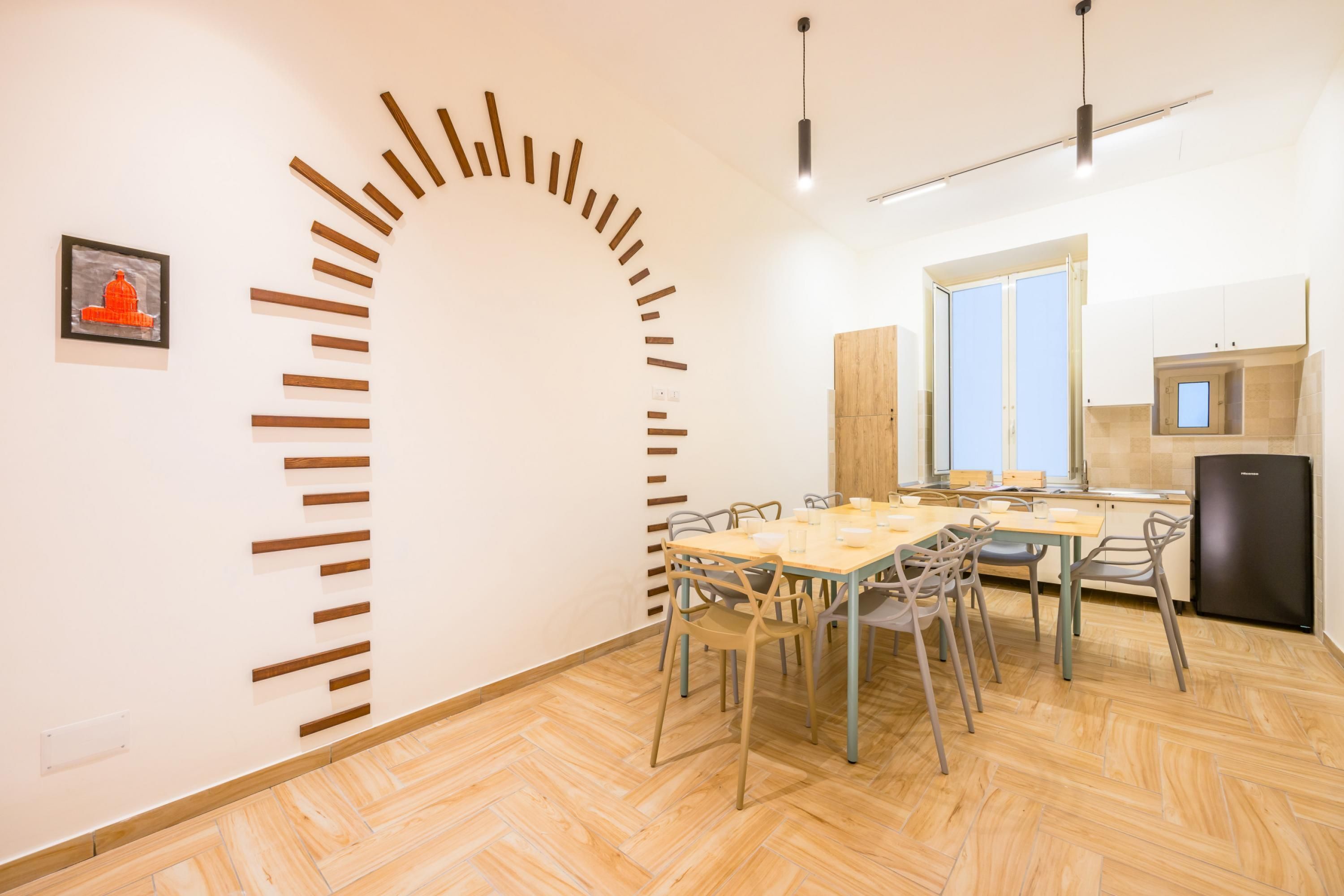 Un espace repas doté d’un plancher en bois, d’une longue table avec des chaises, d’un luminaire moderne et d’un coin cuisine avec des placards et un réfrigérateur.