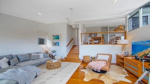 A modern living room with a grey sofa, a round coffee table, and a large rug. There is a staircase leading to an upper level with a view of the sea.