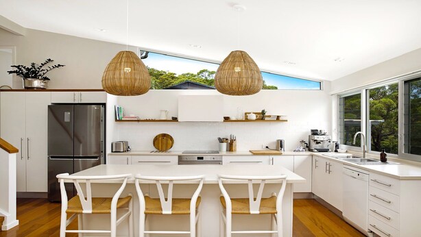 A modern kitchen with white cabinetry, a central island with bar stools, a built-in oven, and a large window offering a view of greenery.