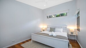 A modern bedroom with a white bed, two bedside lamps, a small table with a vase, and a ceiling fan.