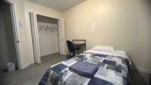A bedroom with a bed, a desk, and a chair. There is a door leading to a closet with clothes hanging.
