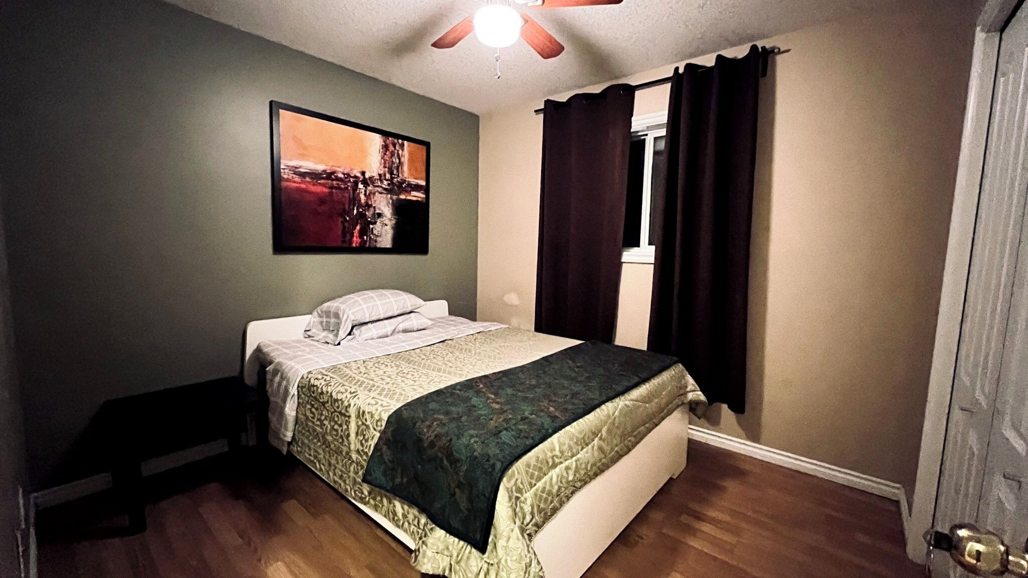A bedroom with a bed, a ceiling fan, a window with curtains, and a painting on the wall.