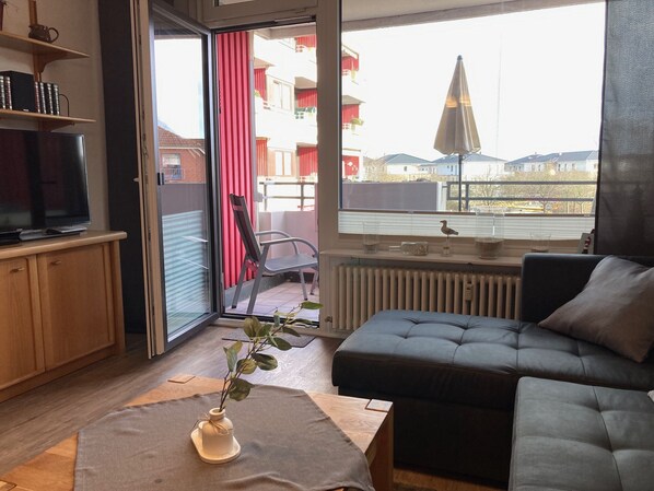 Ein modernes Wohnzimmer mit einer Couch, einem Fernseher auf einem Holzschrank, einer Schiebetür zum Balkon und Blick auf die Stadt.