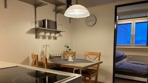 Une cuisine compacte équipée d’une cuisinière, d’un four à micro-ondes et de tasses suspendues. On y trouve une table à manger avec des chaises et une horloge au mur. Une fenêtre donne sur un ciel bleu.