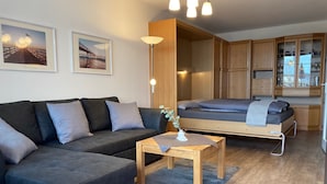 Un salon moderne avec un canapé, une petite table et un lit avec un cadre en bois. On y trouve également des armoires en bois et une lampe.
