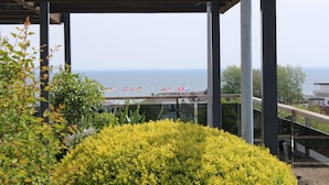 Un espace extérieur abrité, avec vue sur la mer, agrémenté de colonnes en métal, de rampes en verre et de plantes à fleurs jaunes.